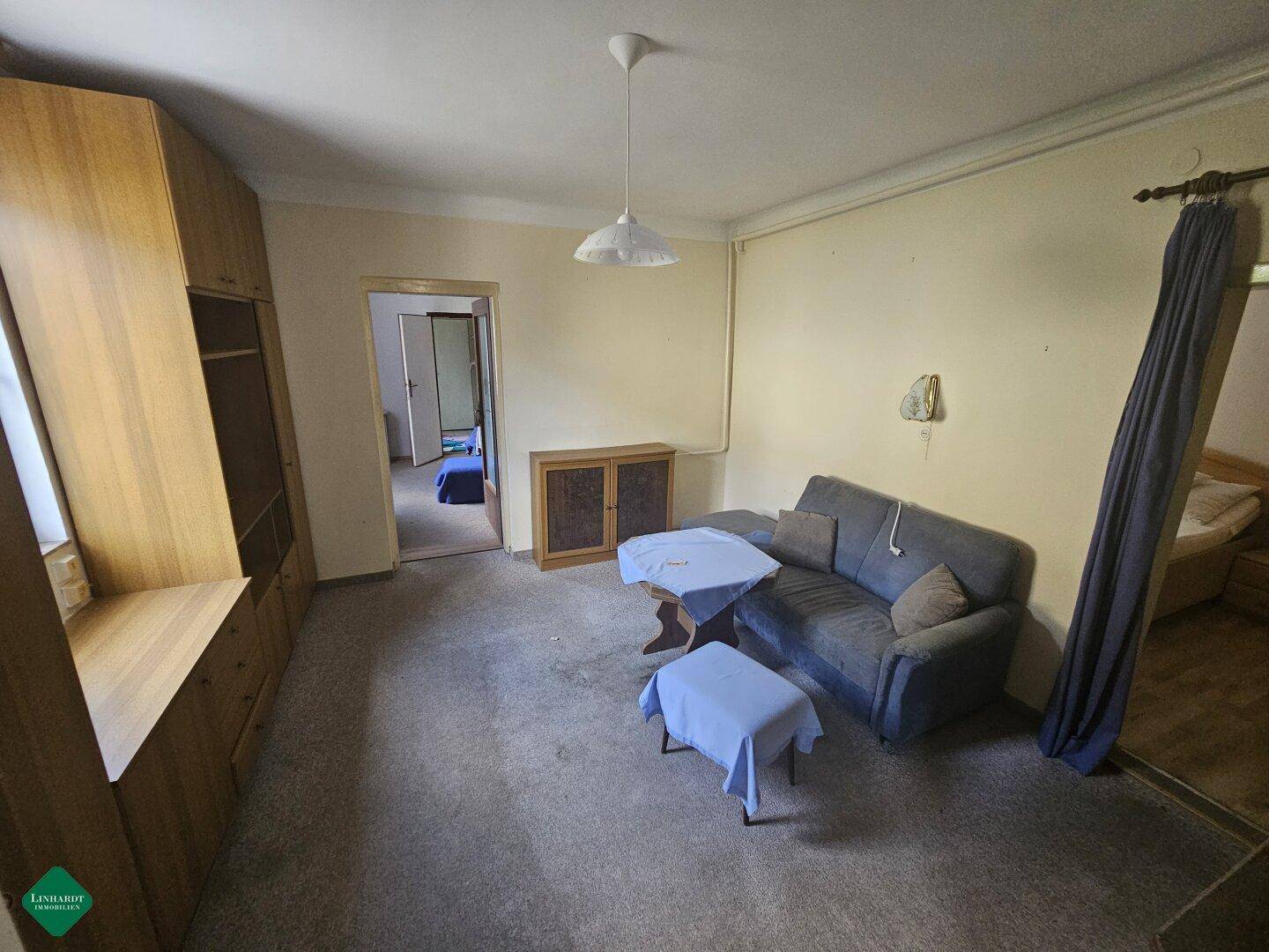 Geräumiges Wohnzimmer mit Teppichboden, Sofa und Holzmöbeln, angrenzend an ein Schlafzimmer.