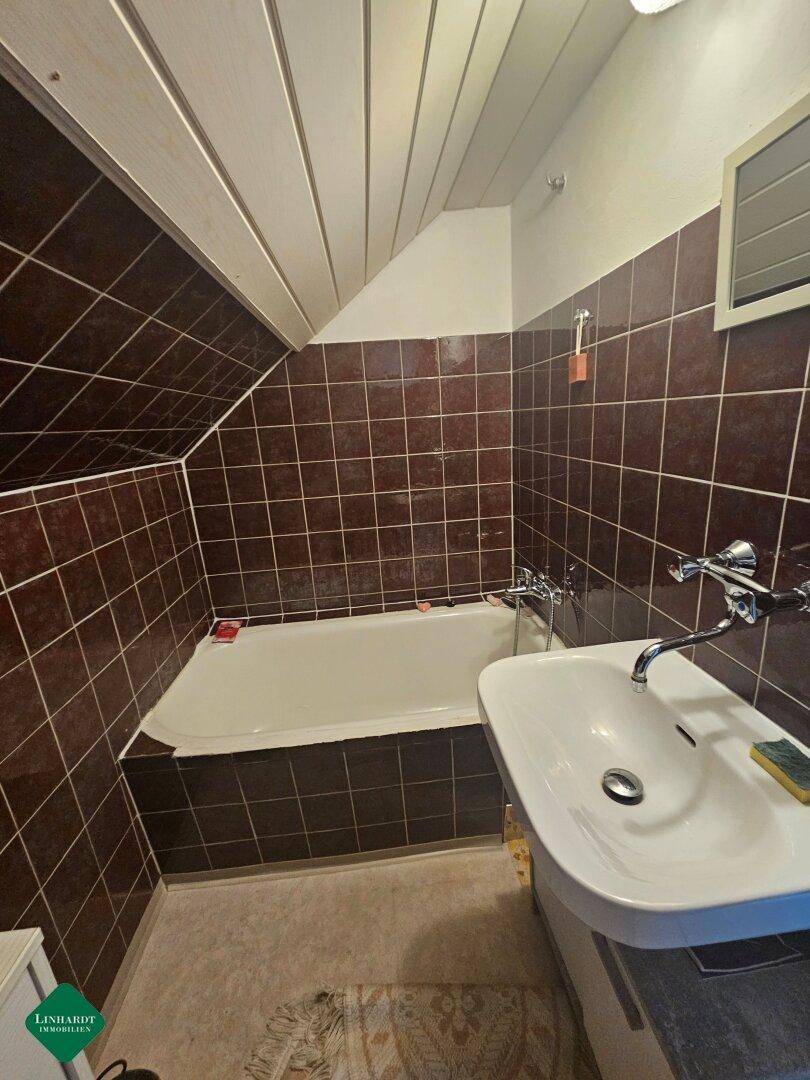 Badezimmer mit braunen Fliesen, Badewanne und Waschbecken unter einer Dachschräge.