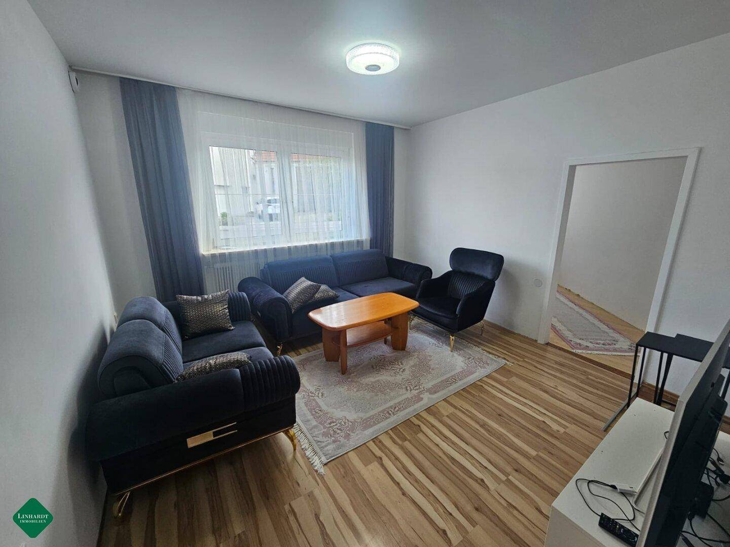 Gemütliches Wohnzimmer mit dunklen Sofas, Holzboden und einem großen Fenster für Tageslicht.