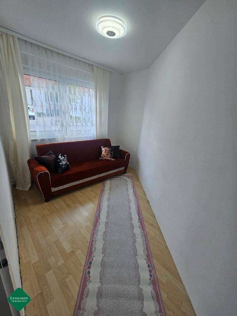 Kleines Zimmer mit rotem Sofa, Laminatboden und Fenster mit hellen Vorhängen.