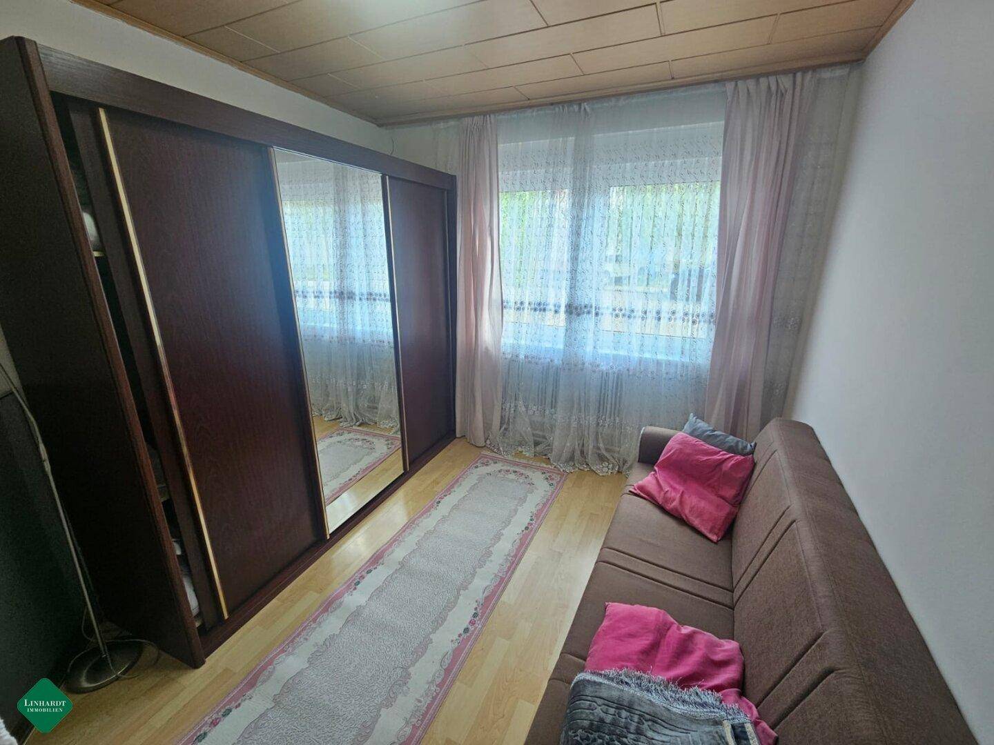 Schlafzimmer mit großem Spiegelschrank, Holzboden und Vorhängen am Fenster.
