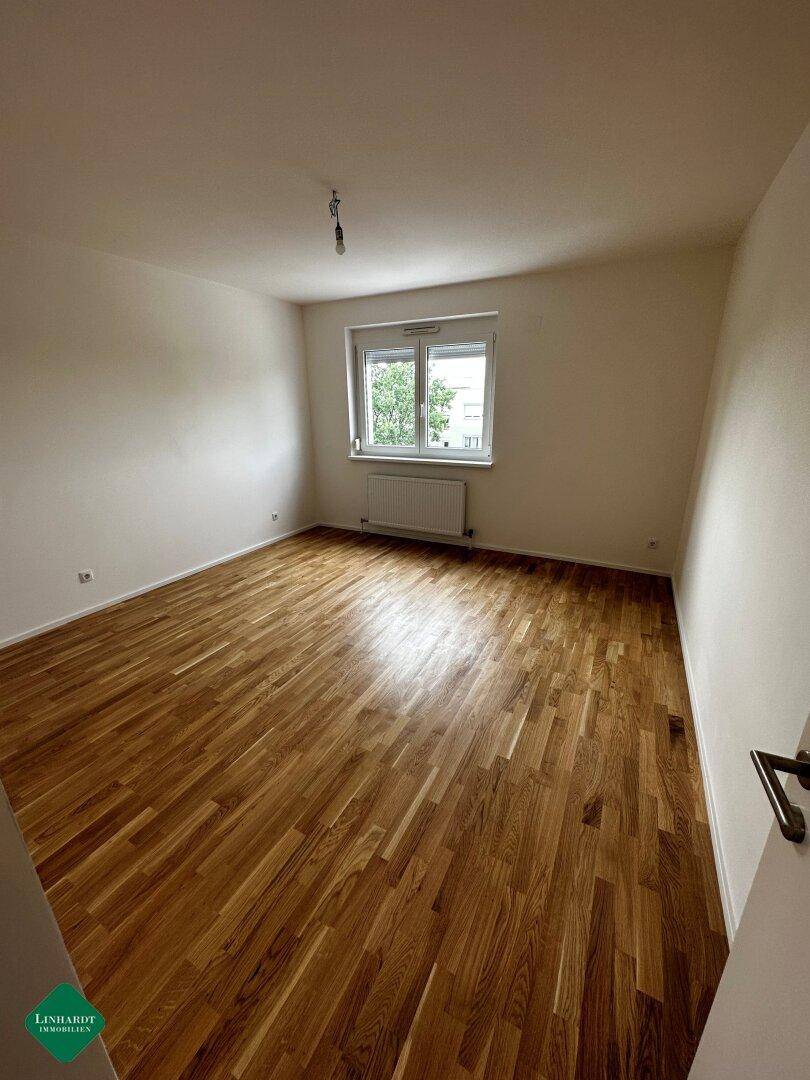 Leeres Schlafzimmer mit warmem Parkettboden und einem Fenster, das viel Tageslicht hereinlässt.