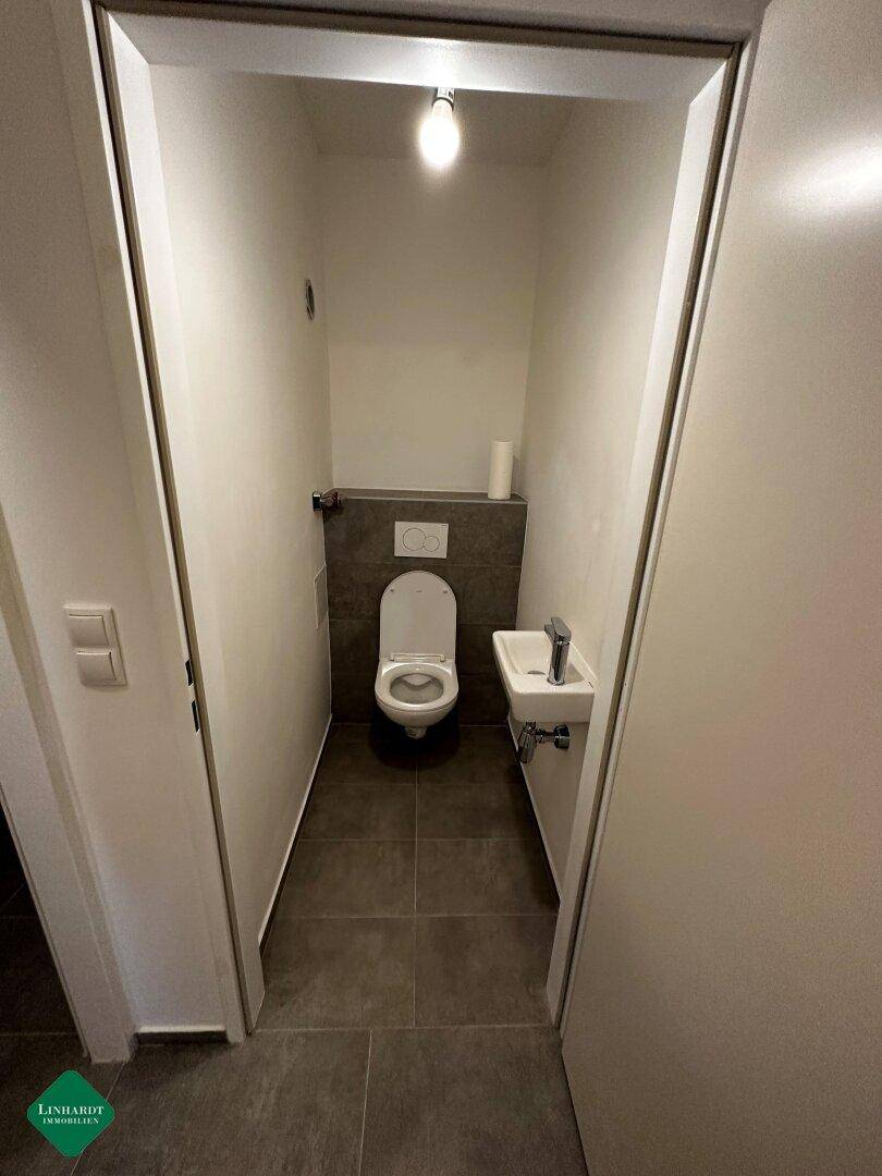 Separates Gäste-WC mit wandhängender Toilette, kleinem Waschbecken und modernen grauen Fliesen.