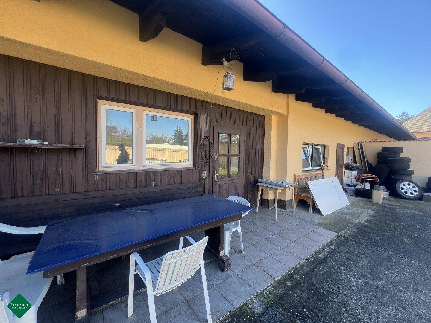 Außenbereich neben dem Gebäude mit Holzwand, gefliestem Boden und provisorischer Sitzgelegenheit.