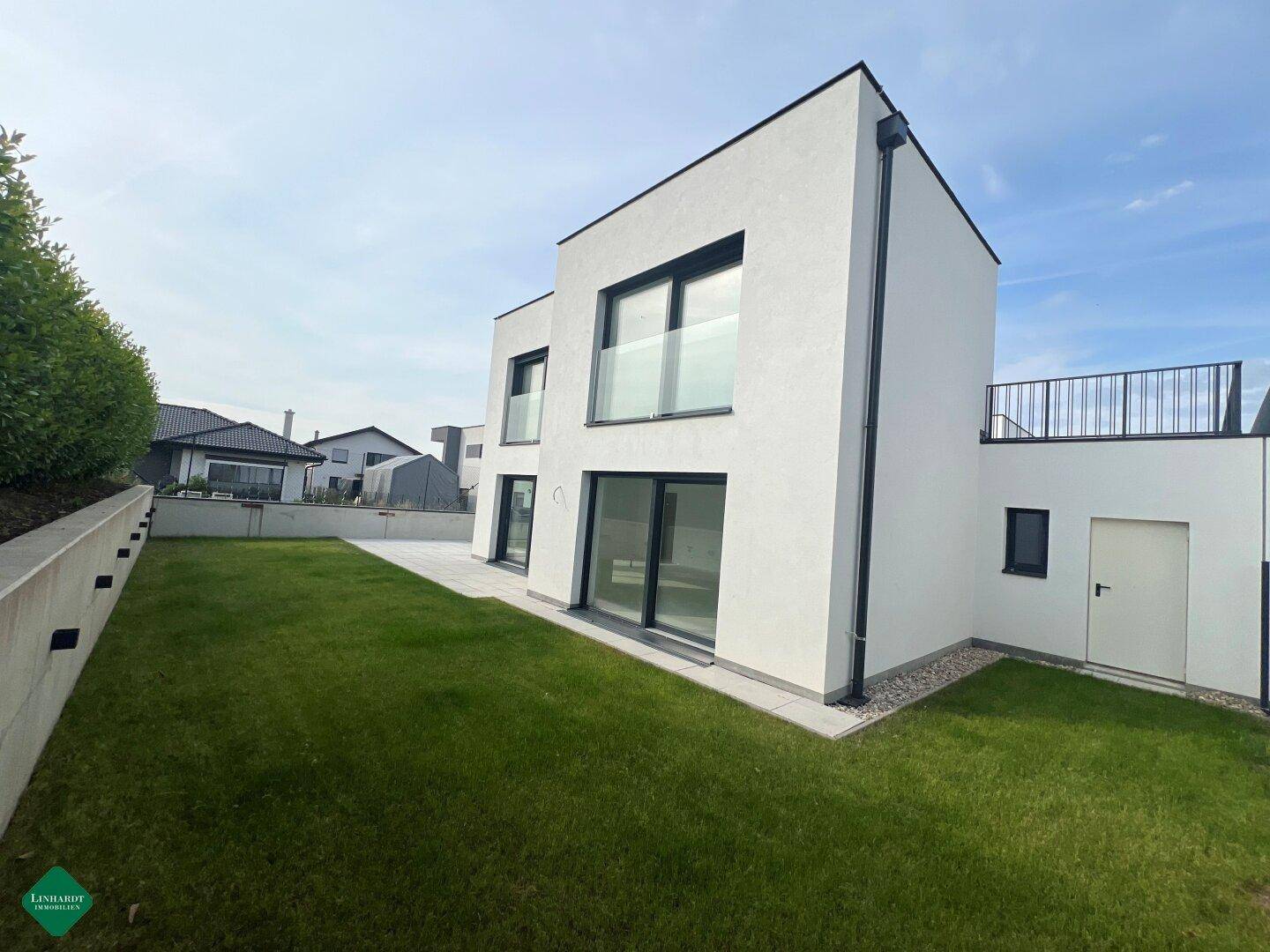 Rückansicht des modernen Hauses mit einer Dachterrasse, Rasenfläche und einer beleuchteten Stützmauer.