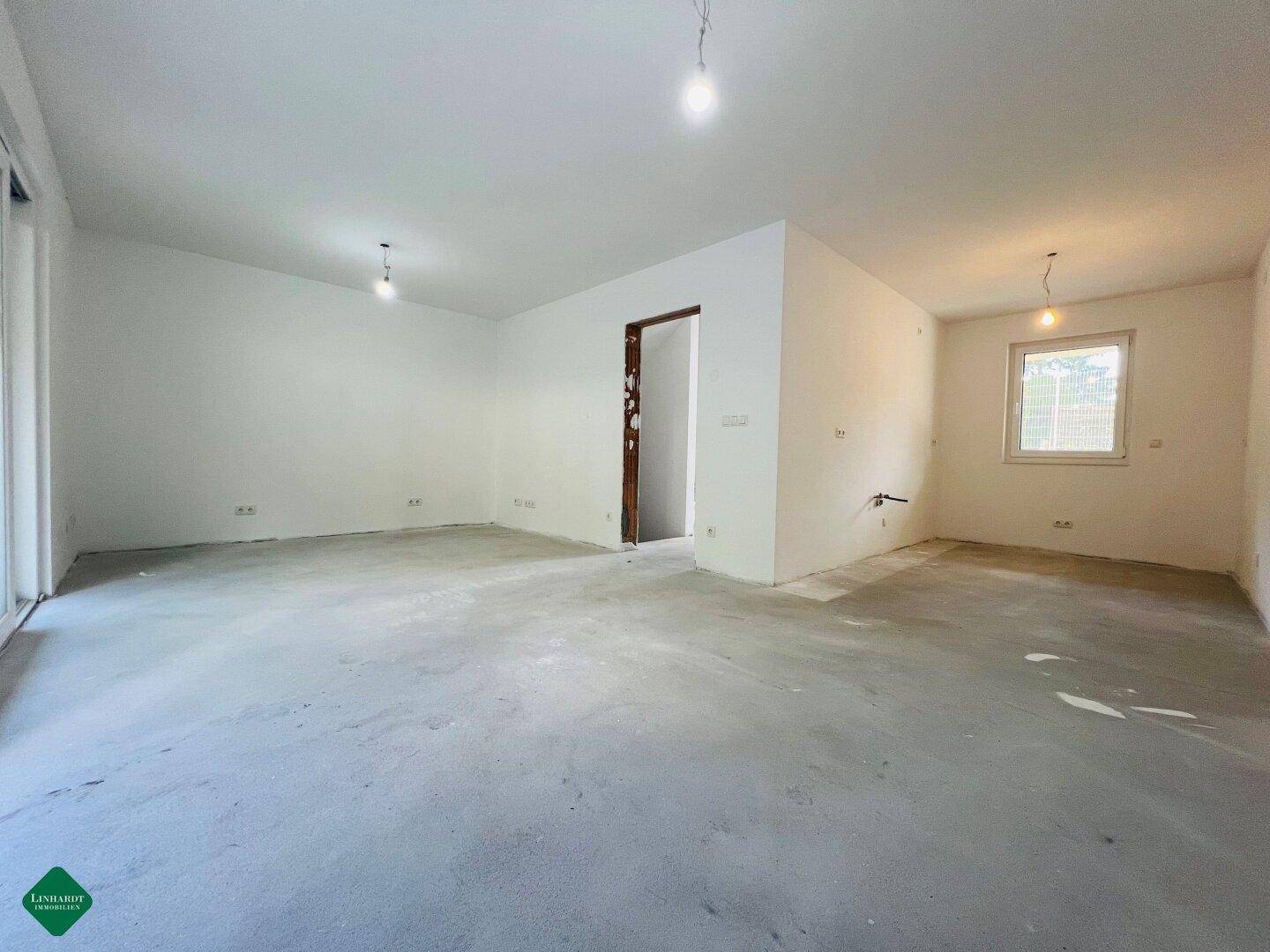 Unfertiger Wohnbereich im Rohbau mit Betonboden, weißen Wänden und Zugang zu einem Außenbereich.