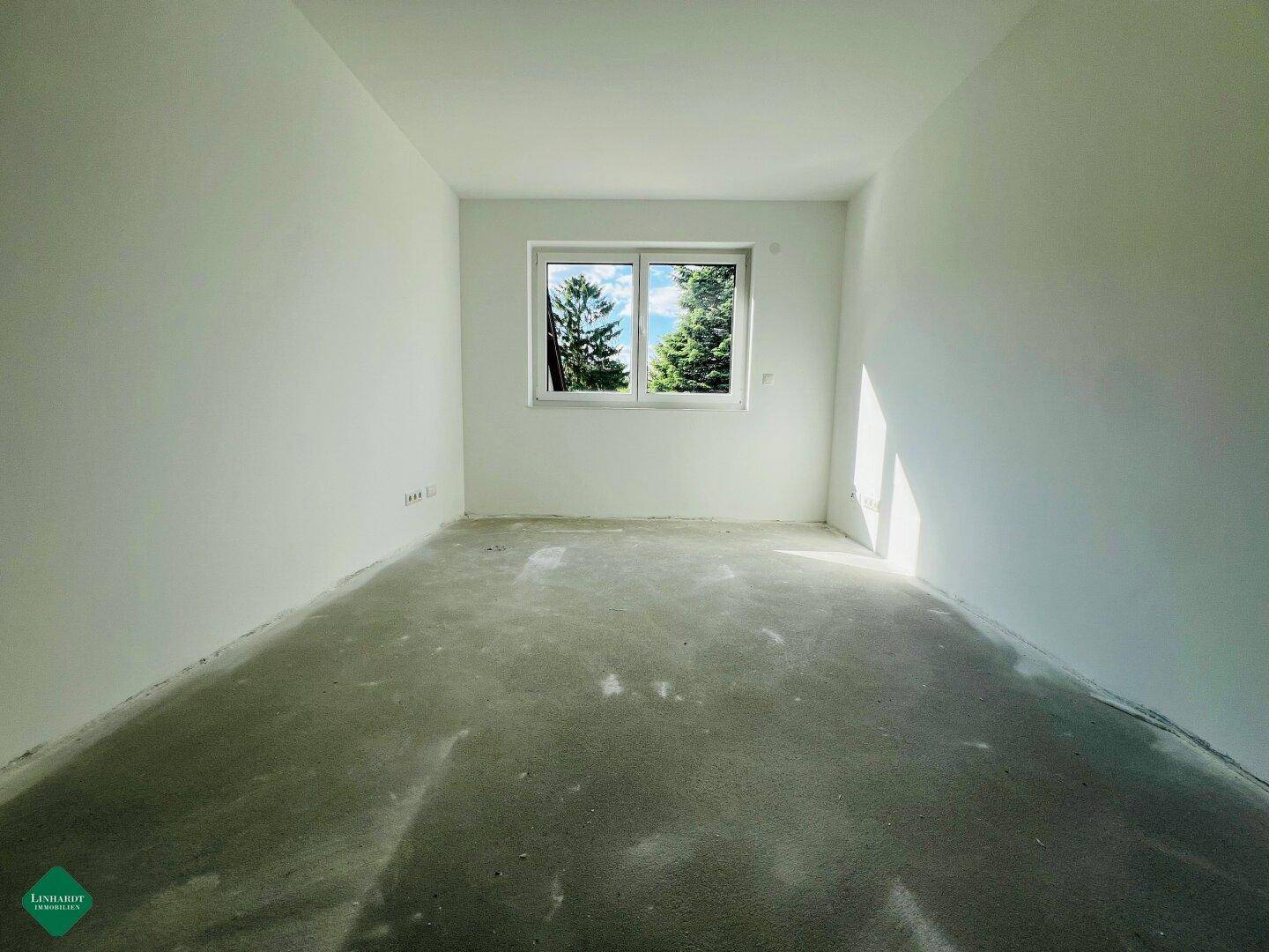 Helles Schlafzimmer im Rohbau mit Betonboden und Blick ins Grüne durch das Fenster.