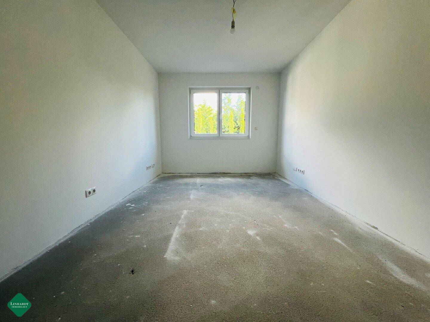 Weiteres Schlafzimmer im Rohbau mit Betonboden, weißen Wänden und Ausblick ins Grüne.