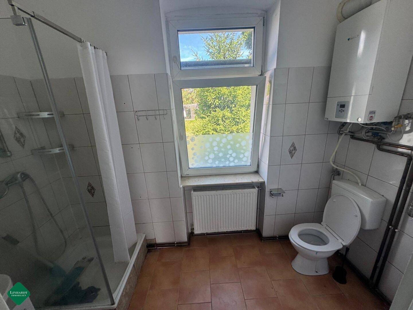 Helles Badezimmer mit Badewanne, WC und Gastherme, gefliest in Weiß und Braun.