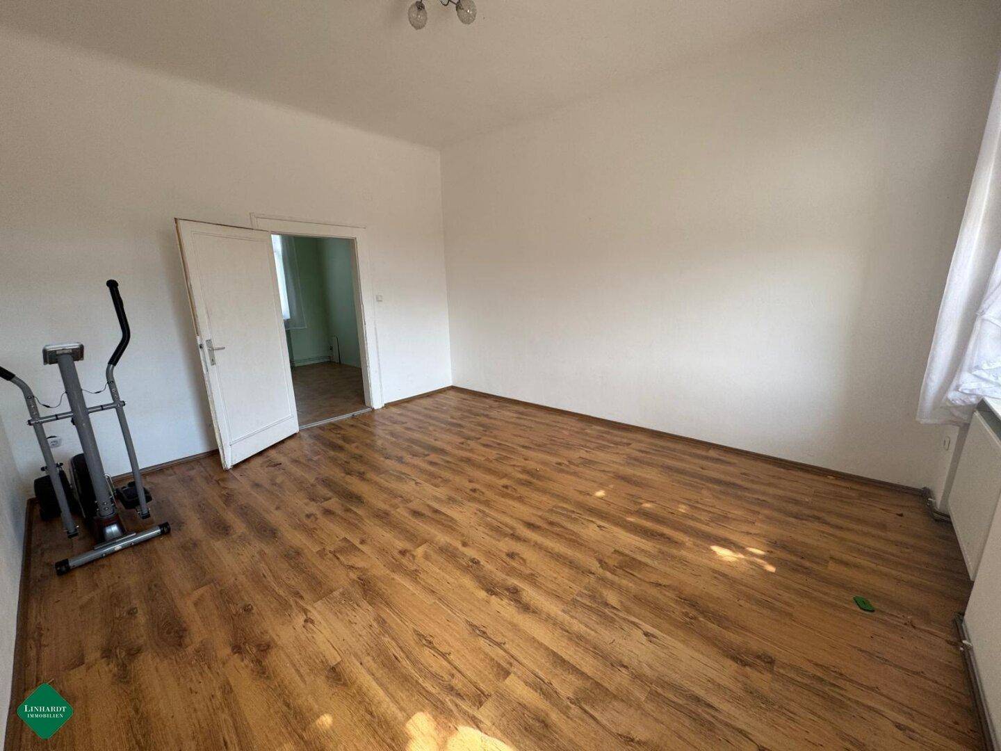 Geräumiges Schlafzimmer mit Laminatboden und weiß gestrichenen Wänden, mit Zugang zu einem weiteren Raum.