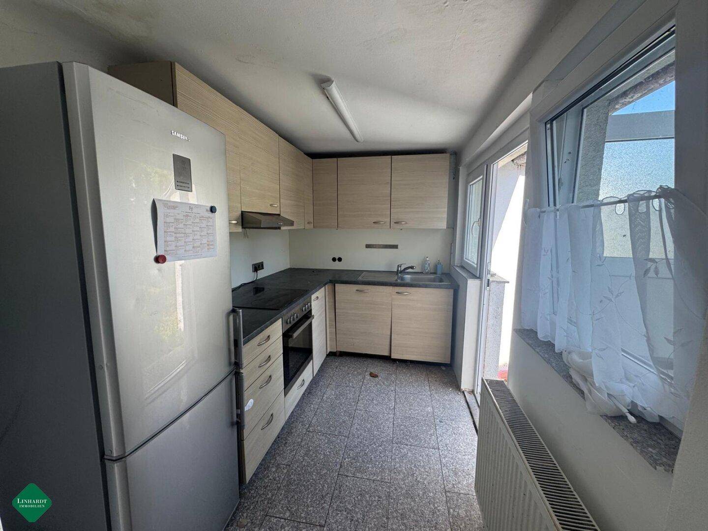Funktionale Küche mit hellen Holzschränken, Kühlschrank und Herd, gefliestem Boden und Fenster.