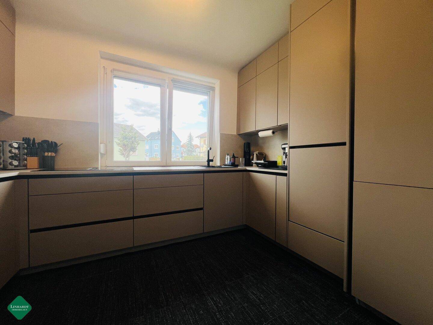Moderne Einbauküche mit beigen Schränken, dunklem Boden und großem Fenster.