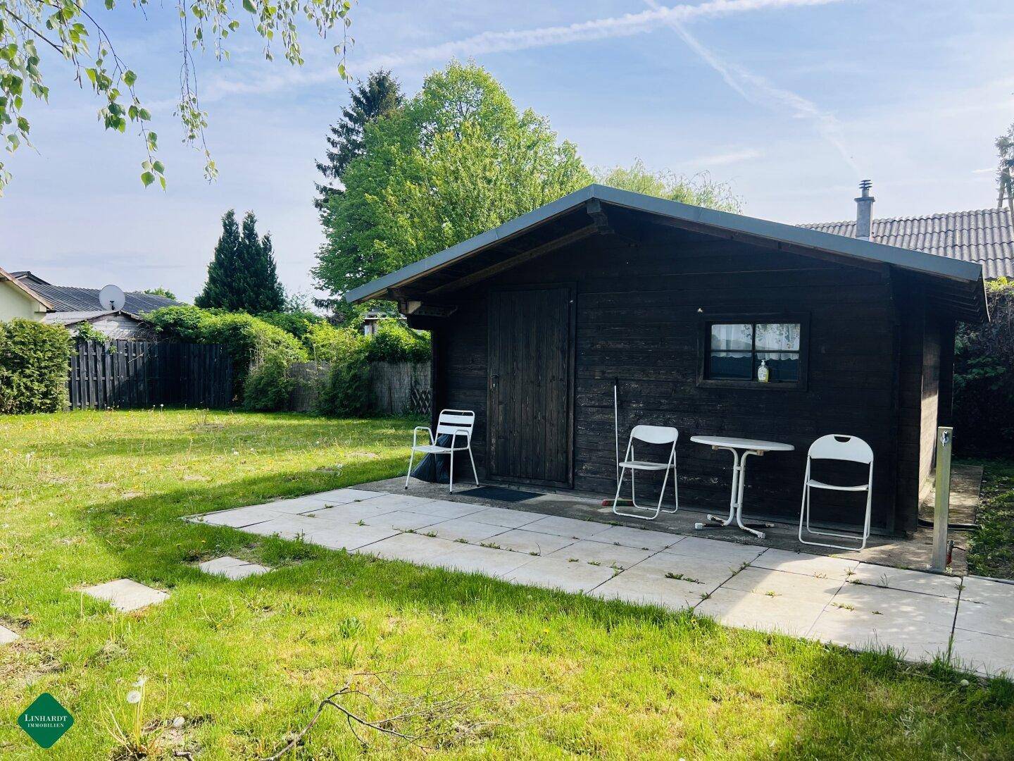 Rustikales Holzhaus mit kleiner gepflasterter Terrasse und Gartenmöbeln im Grünen.