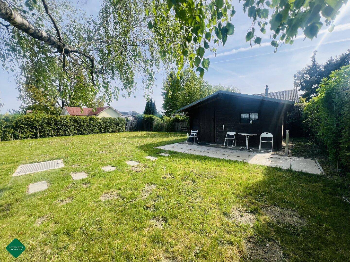 Sonniger Garten mit Rasenfläche, Trittsteinen und einem kleinen Holzhaus mit Sitzgelegenheit.