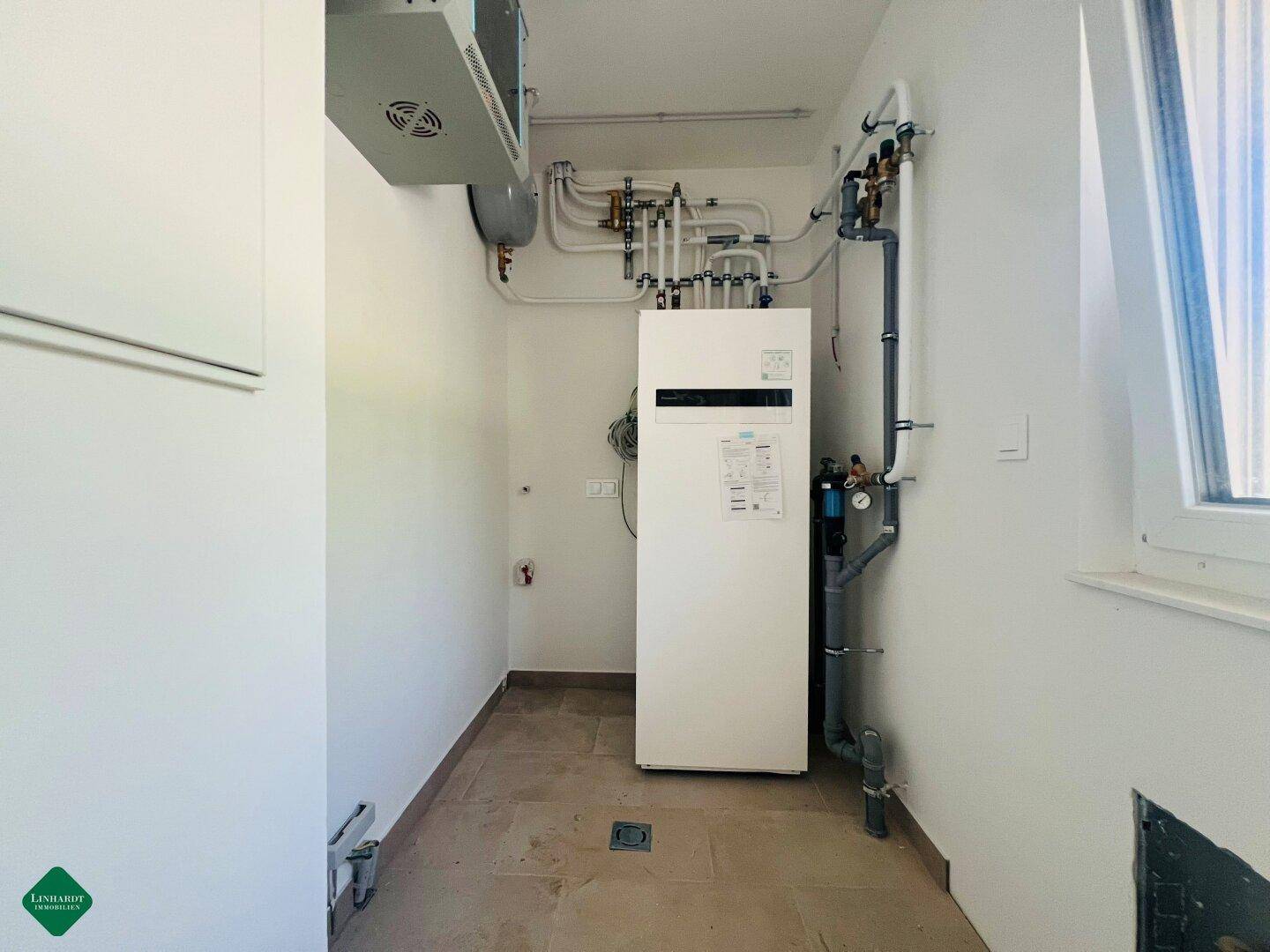 Technikraum mit moderner Wärmepumpe, Rohrleitungen und einem Fenster für Belüftung.