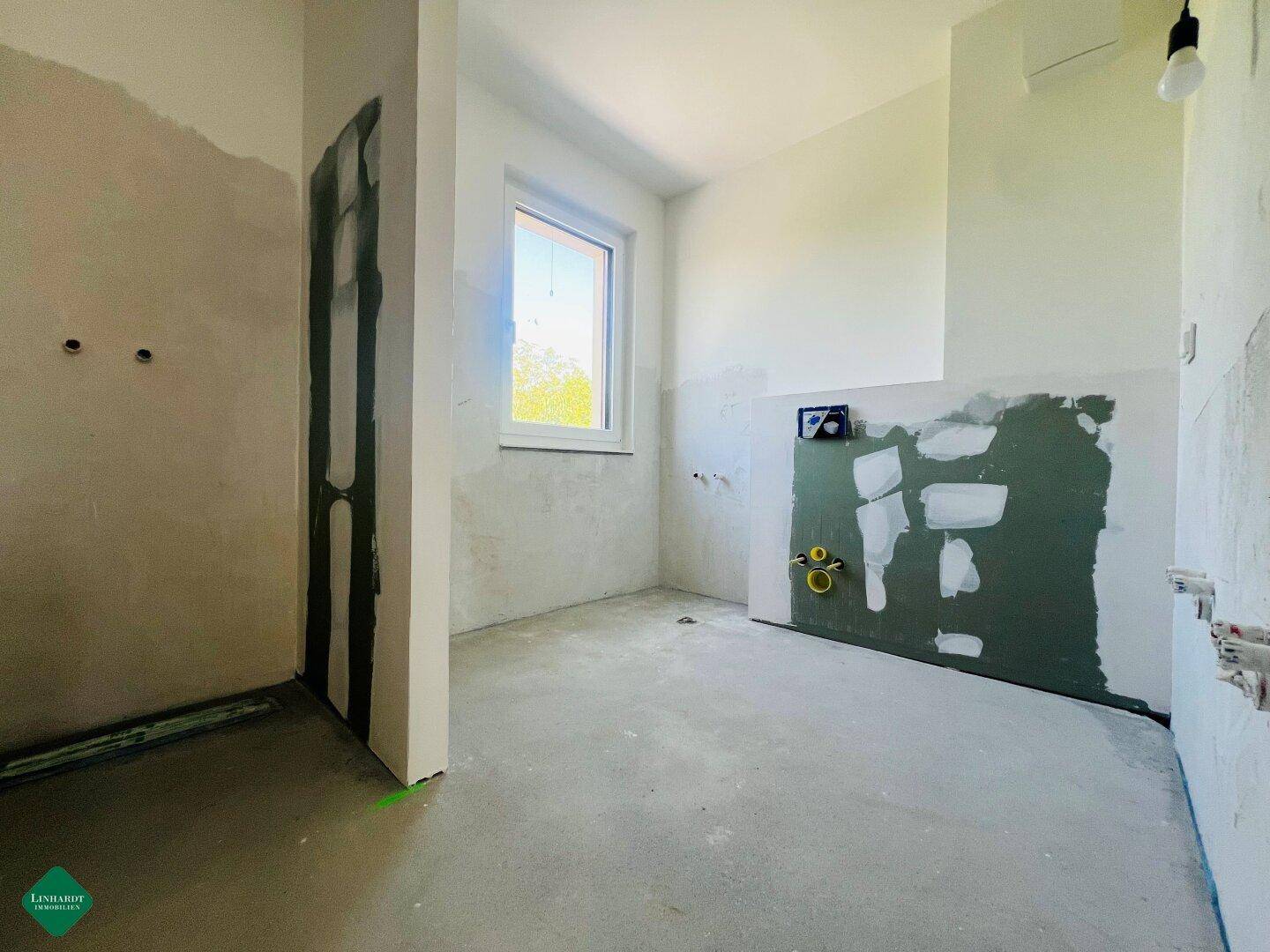 Unfertiges Badezimmer mit Fenster und vorbereiteten Anschlüssen für Sanitärinstallationen.