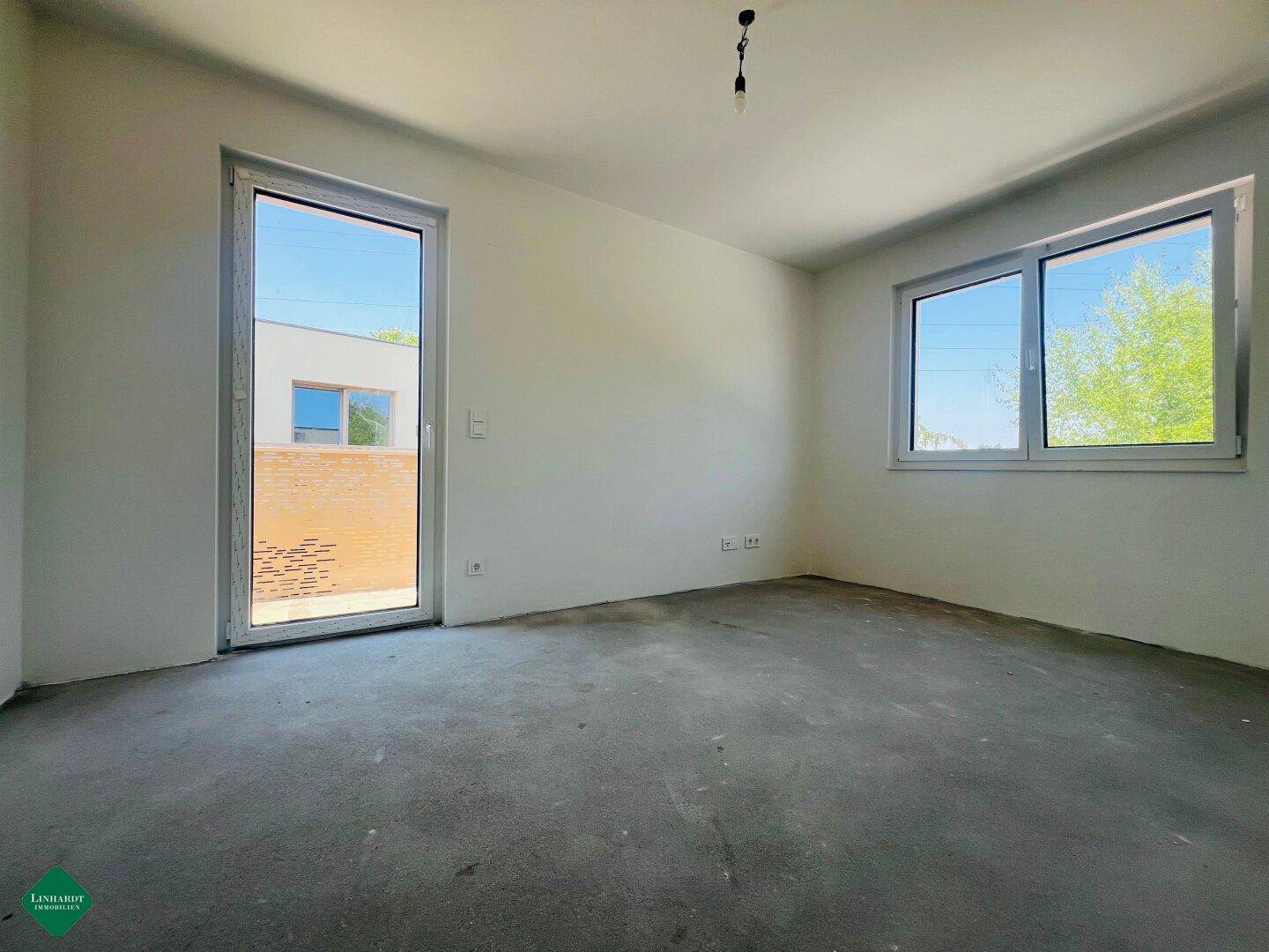Helles, unmöbliertes Zimmer im Neubau mit großen Fenstern und Blick ins Freie.