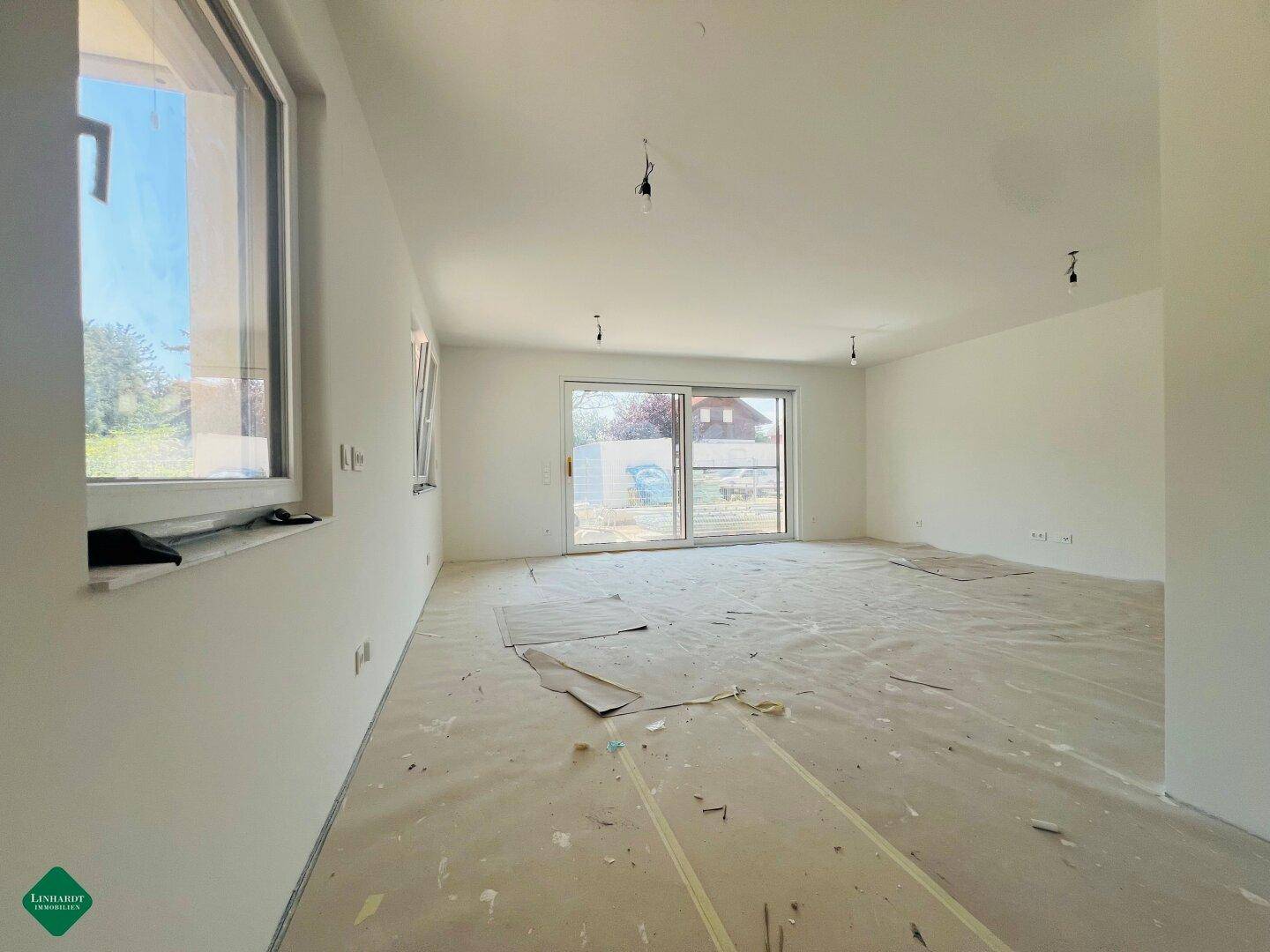 Leerer Raum im Rohbau mit großen Fenstern und Schiebetür zum Außenbereich, viel Tageslicht.
