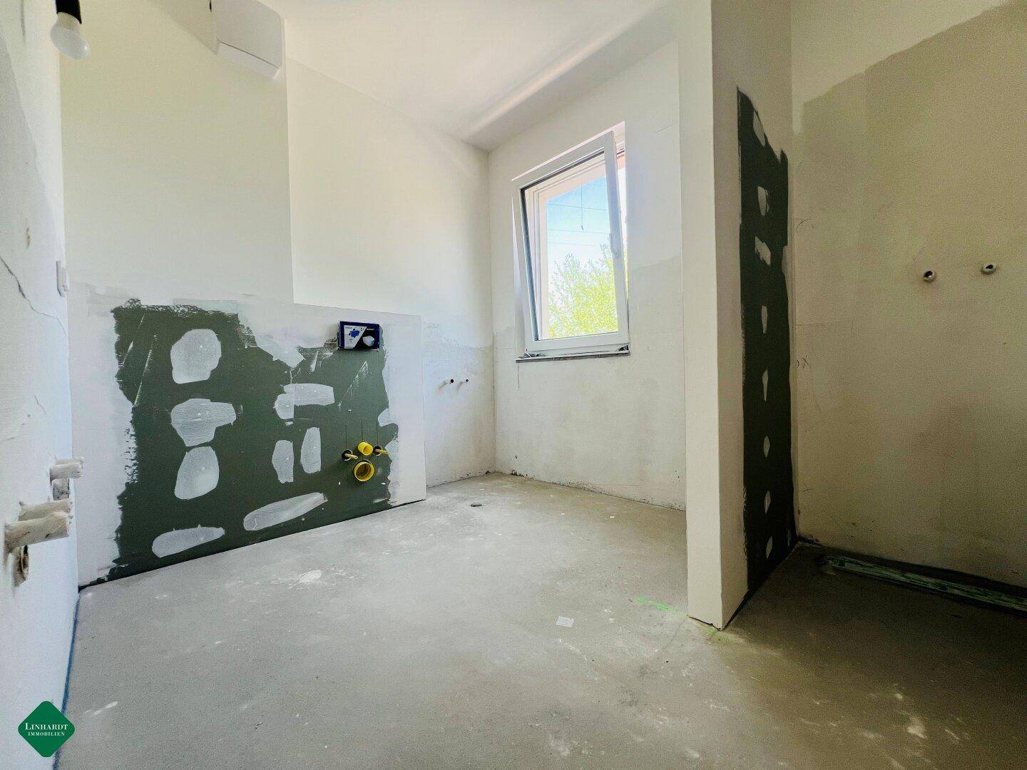 Rohbauküche mit vorbereiteten Anschlüssen für Spüle und Geräte sowie einem Fenster für Tageslicht.