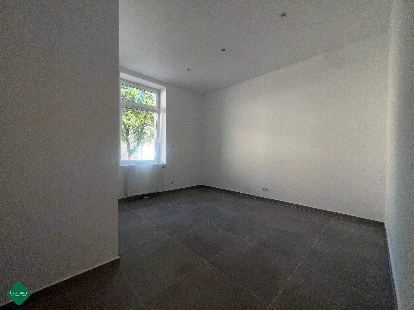 Leerer Raum mit großem Fenster, das viel Tageslicht hereinlässt, und grauen Bodenfliesen.