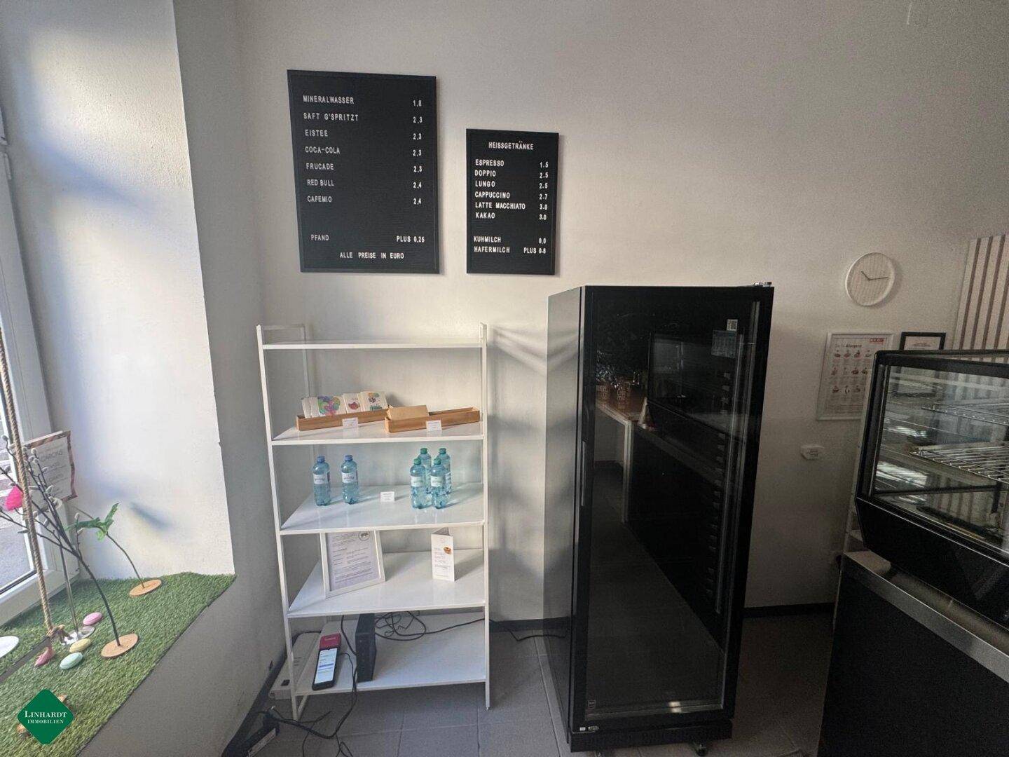 Bereich eines Cafés mit Getränkekühlschrank, weißen Regalen und Menütafeln an der Wand.