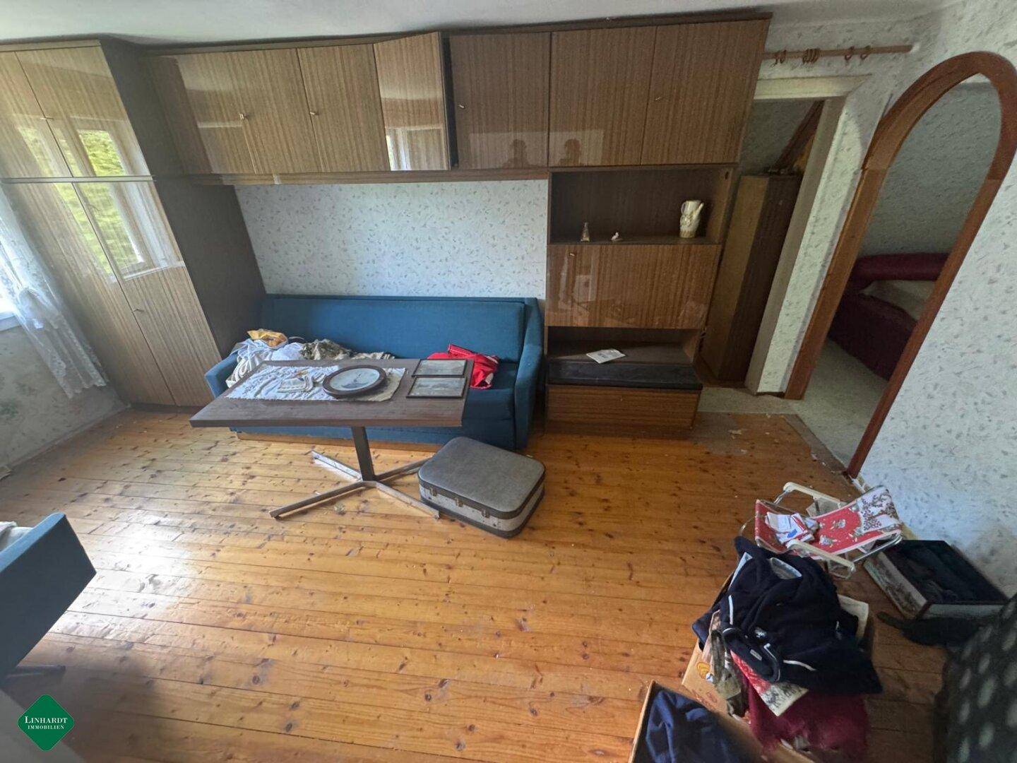 Wohnzimmer mit Holzfußboden, blauer Couch und älteren Einbauschränken, renovierungsbedürftig.