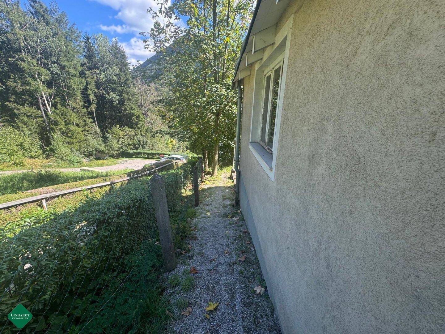 Schmaler Weg entlang der Hauswand mit Hecke und Blick auf die umliegende Natur.