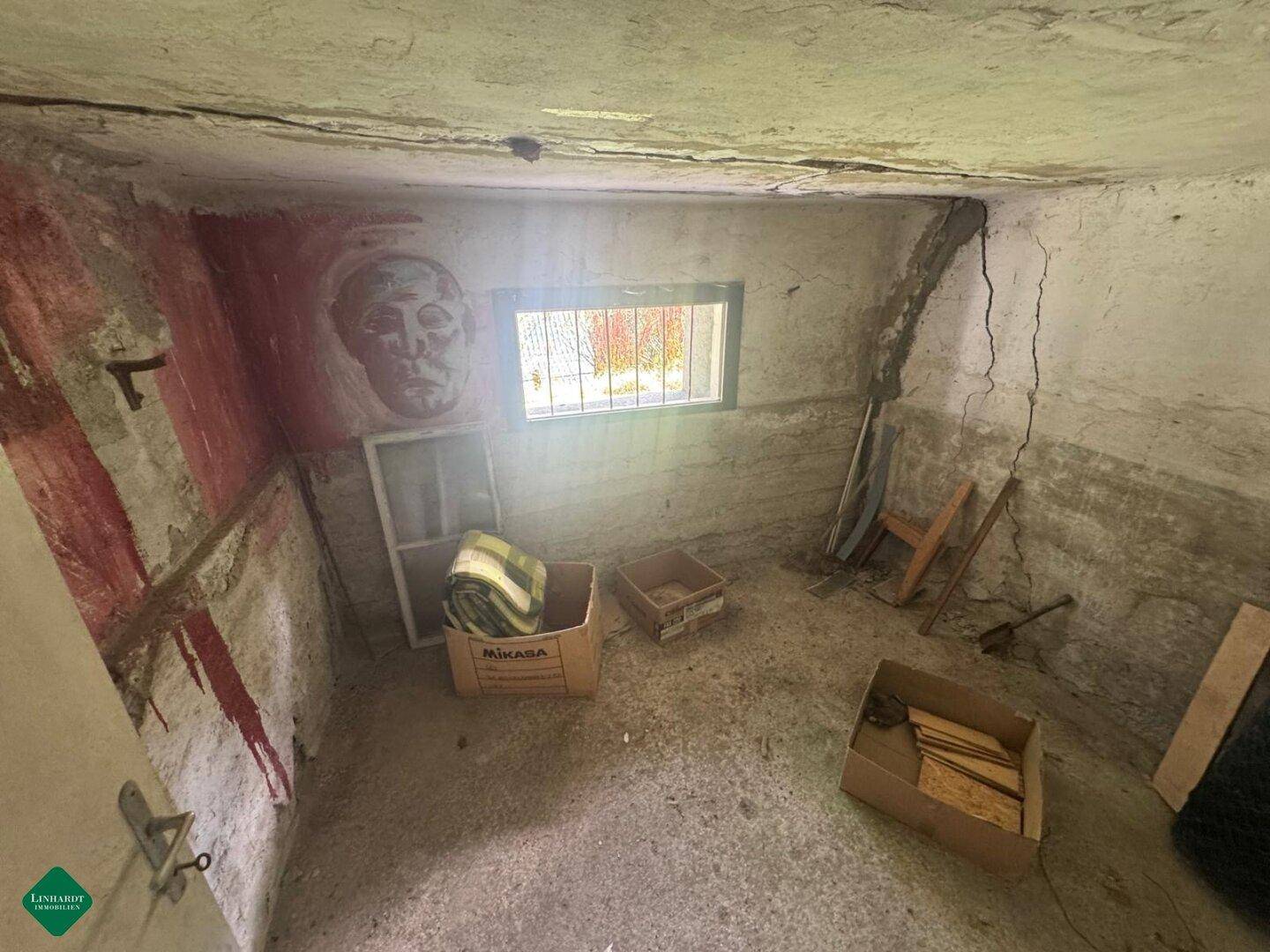 Kellerraum mit Betonboden, vergittertem Fenster und Wandmalerei, in sanierungsbedürftigem Zustand.