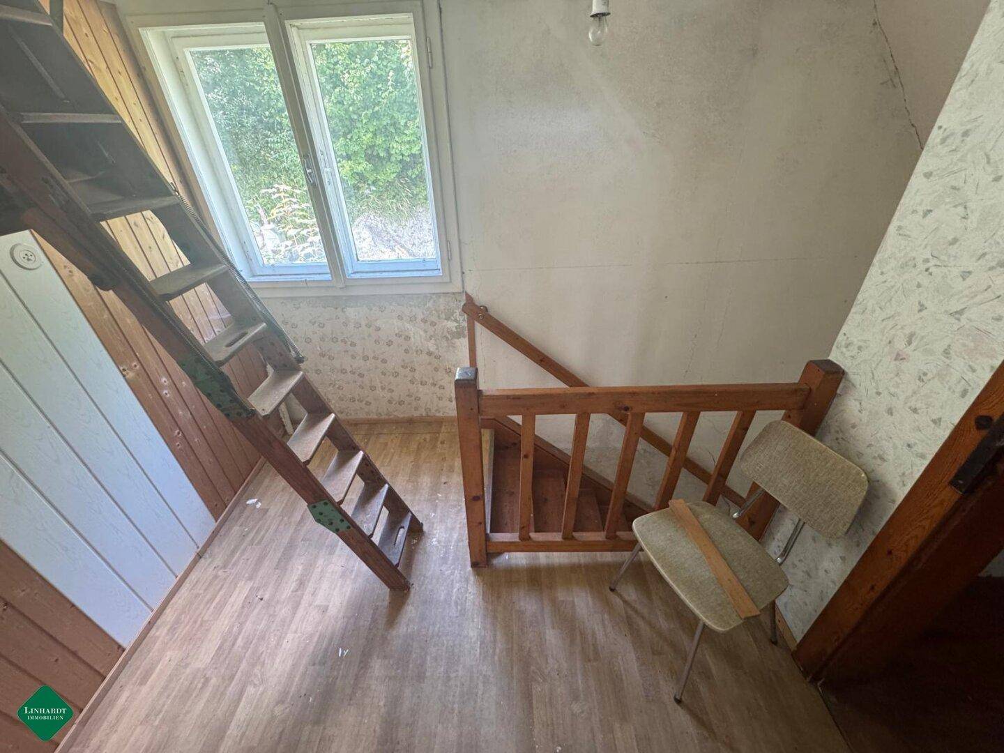 Flur mit Holztreppe, Fenster und Blick ins Grüne, renovierungsbedürftig.
