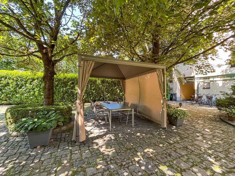 Gemütlicher Sitzbereich im Innenhof unter einem Pavillon, umgeben von Bäumen und Grün.