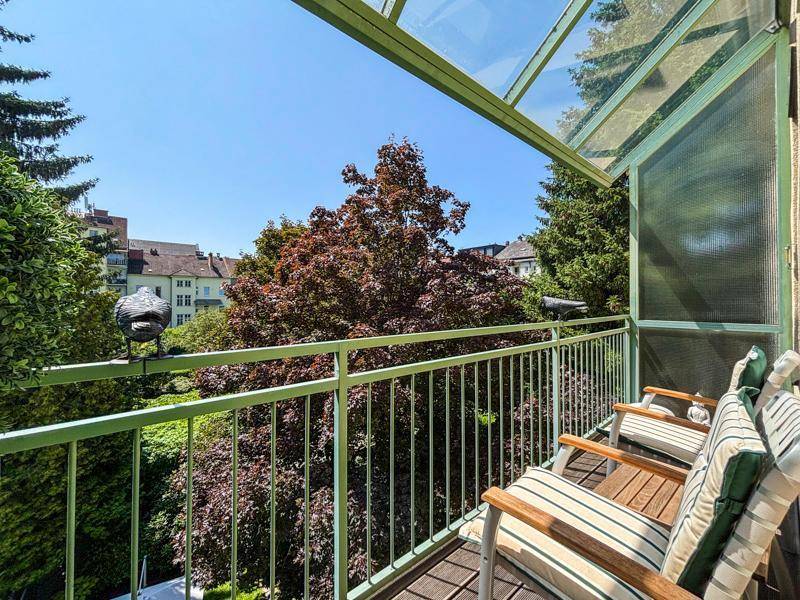 Gemütlicher Balkon mit Sitzgelegenheiten und Blick auf grüne Bäume unter blauem Himmel.