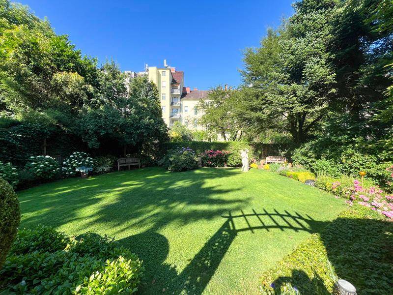 Gepflegter, weitläufiger Garten mit Rasenfläche, Blumenbeeten und hohen Bäumen im Sonnenlicht.