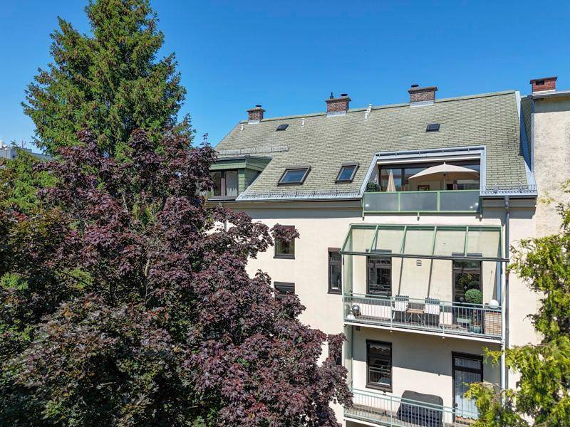 Rückansicht eines Gebäudes mit mehreren Balkonen und einer Dachterrasse, umgeben von Bäumen.