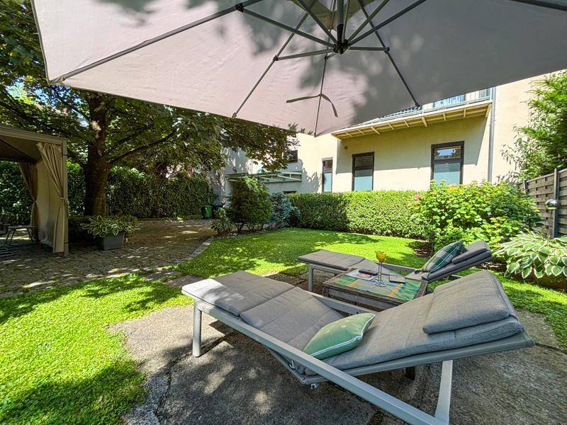 Entspannungsbereich im Garten mit Liegestühlen und Sonnenschirm, umgeben von Rasen und Hecken.