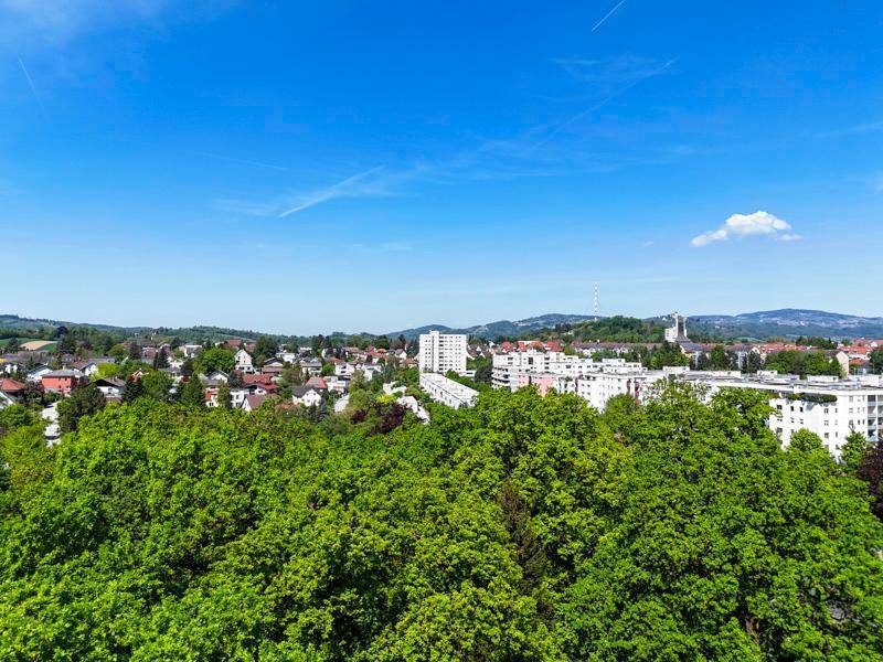 Panoramablick über eine Stadtlandschaft, umgeben von üppigen grünen Bäumen und sanften Hügeln.