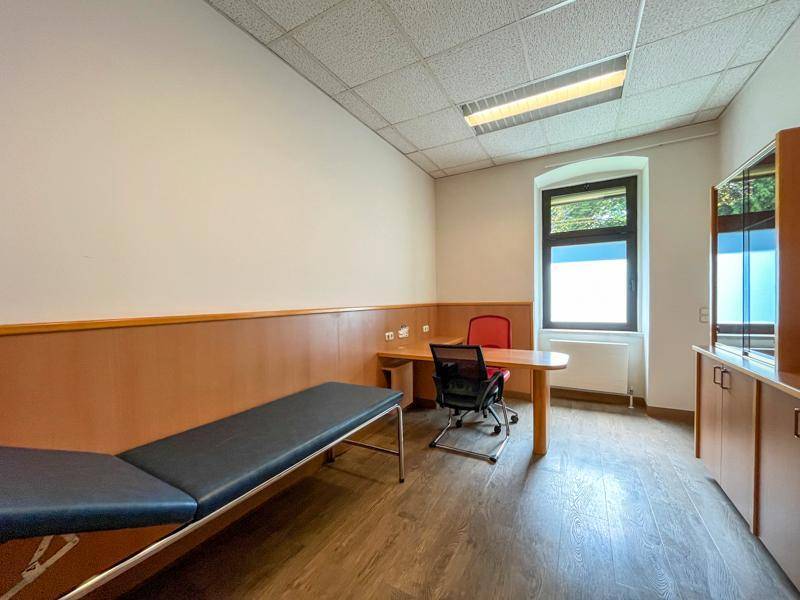 Helles Behandlungs- oder Beratungszimmer mit Liege, Schreibtisch und Laminatboden.