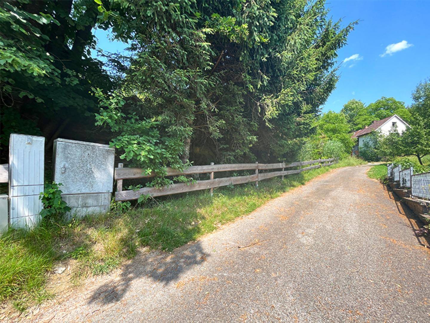Schotterweg führt entlang eines Holzzauns und alter Stromkästen, mit einem Haus im Grünen am Horizont.