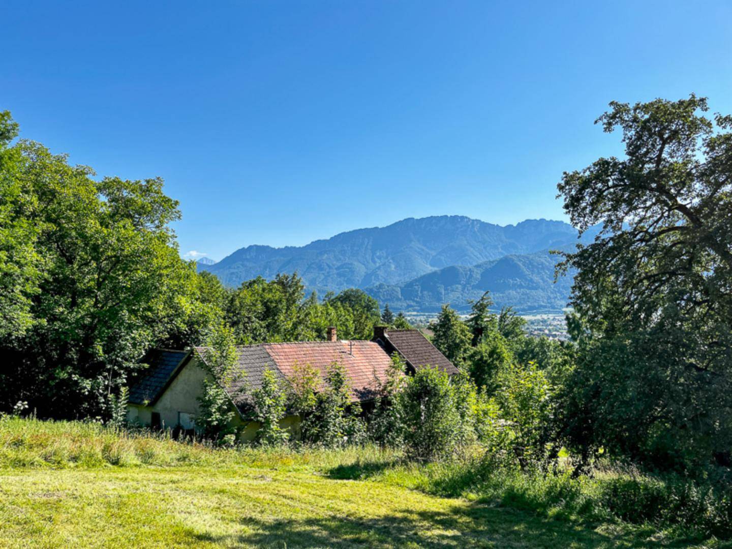 Idyllisches Haus mit Ziegeldach, umgeben von Bäumen und einem beeindruckenden Bergpanorama in der Ferne.