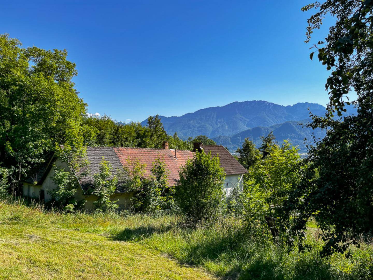 Altes Haus mit Ziegeldach, umgeben von üppigem Grün und einem malerischen Bergpanorama im Hintergrund.