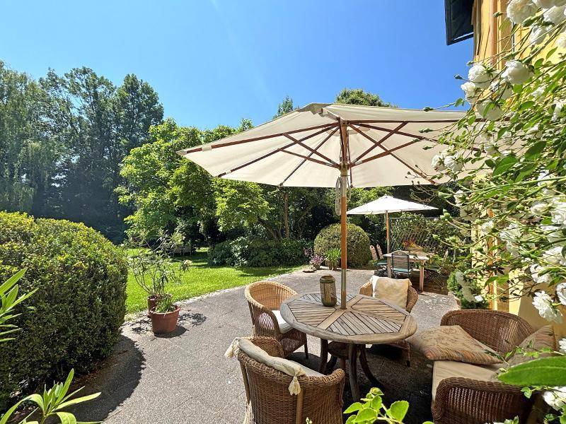 Sonnige Terrasse mit Korbmöbeln und großem Sonnenschirm, umgeben von üppigem Grün und blühenden Pflanzen.