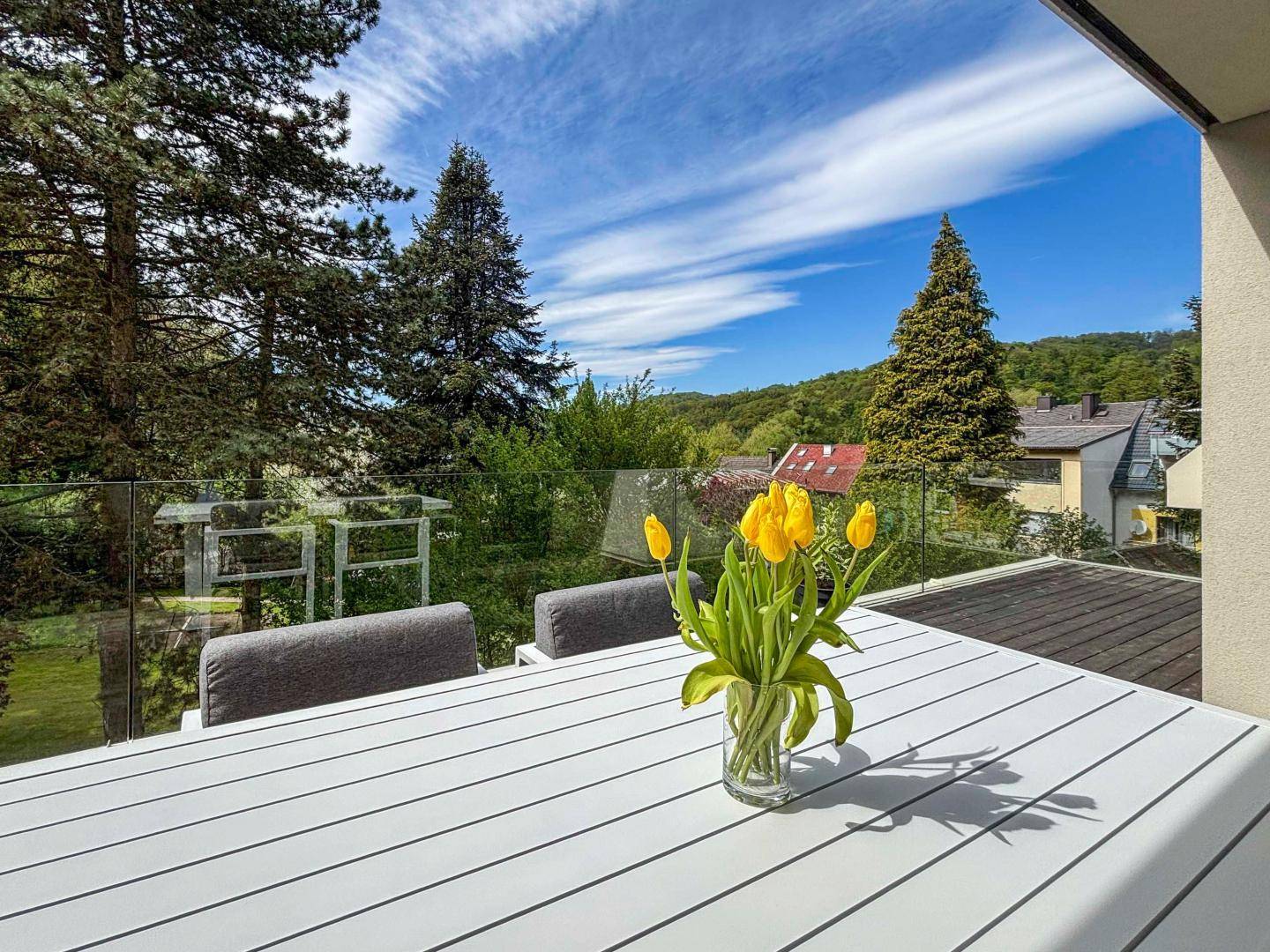 Sonniger Balkon mit Esstisch, Glasgeländer und weitem Blick ins Grüne.