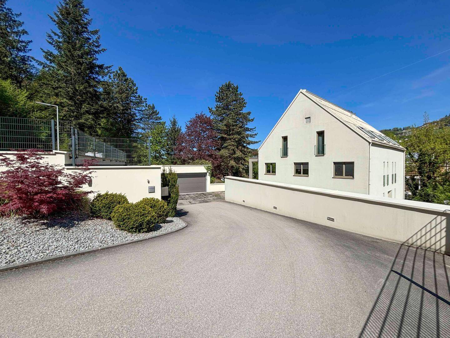 Modernes Einfamilienhaus mit heller Fassade, Zufahrt und gepflegter Außenanlage unter blauem Himmel.