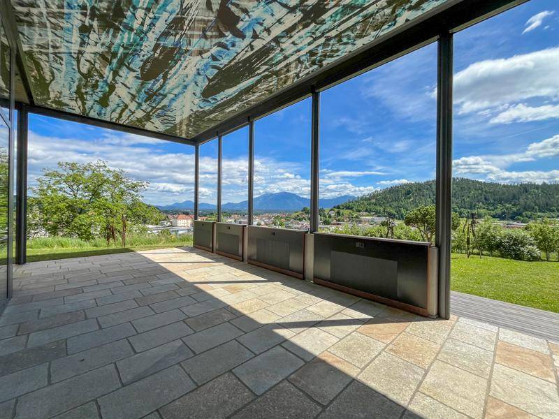 Überdachte Außenterrasse mit Steinfliesenboden, großen Fenstern und weitem Blick auf Landschaft und Berge.