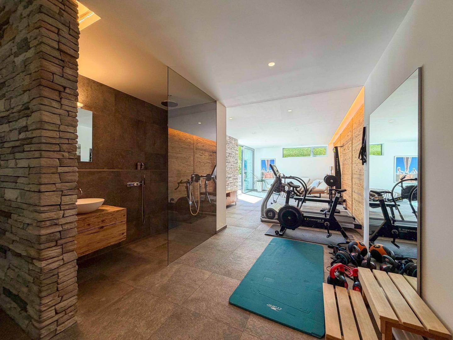 Fitnessraum mit modernen Trainingsgeräten, Steinwand und integriertem Duschbereich.