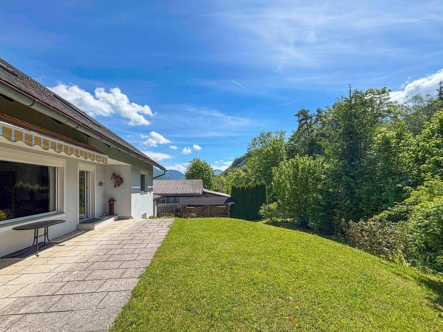 Gepflasterte Terrasse neben dem Haus mit Rasenfläche und weitem Blick in die Natur.