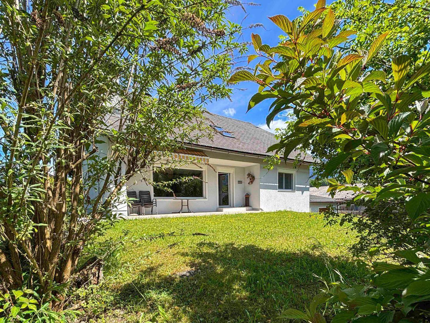 Hausansicht durch grüne Sträucher, mit kleiner Terrasse und Rasenfläche unter blauem Himmel.