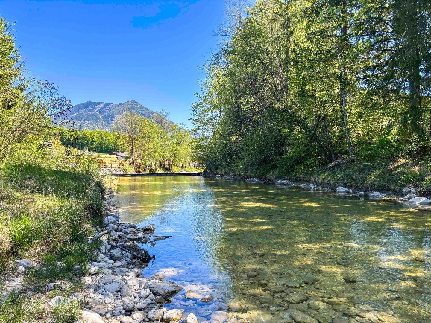 Klarer Flusslauf mit felsigem Ufer und bewaldeten Bergen im Hintergrund an einem sonnigen Tag.