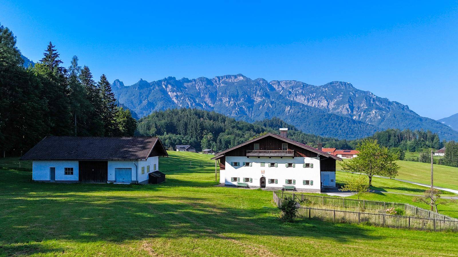Anwesen mit Haupthaus und Nebengebäude, eingebettet in eine weite grüne Landschaft mit majestätischen Bergen im Hintergrund.