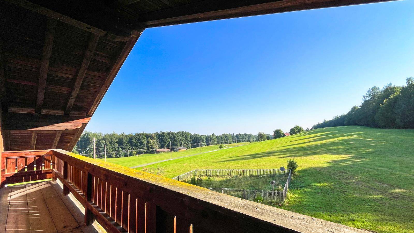 Geräumiger Holzbalkon mit weitem Blick über grüne Wiesen und Wälder unter strahlend blauem Himmel.
