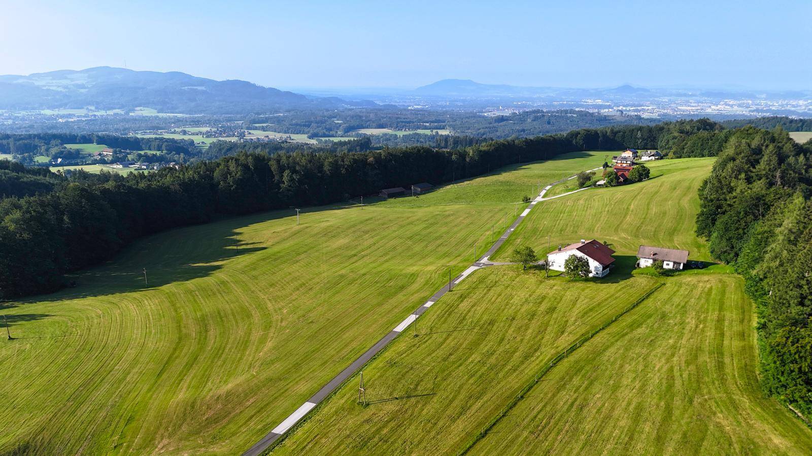 Panoramische Luftaufnahme einer weitläufigen grünen Hügellandschaft mit vereinzelten Häusern und Fernblick.